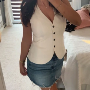 Vit figursydd väst med knappar - Snygg vit väst med v-ringad hals och fyra svarta knappar framtill. Figursydd modell som ger en clean och stilren look. Perfekt att styla över en t-shirt eller direkt på huden för en trendig vibe.