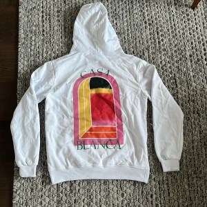 Ljusblå hoodie från Casablanca - Ljusblå hoodie från Casablanca med färgglatt grafiskt tryck i rosa, gult, rött och svart på både bröstet och ryggen. Klassisk huva och känguruficka framtill. Mjuk bomullskänsla och relaxed fit, perfekt för en chill look. Passar M