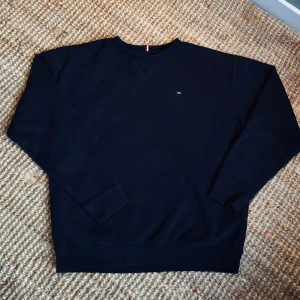 Mörkblå sweatshirt från Tommy Hilfiger - Klassisk mörkblå sweatshirt från Tommy Hilfiger med rund halsringning och diskret broderad logga på bröstet. Tröjan har ribbade muddar vid ärmslut och nederkant samt en snygg detalj med rödvit rand i nacken. Perfekt för en clean och stilren look.