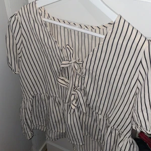 Randig blus med knyt och volang - Säljer en vit blus med svarta ränder och knyt framtill. Blusen har korta ärmar, v-ringning och volang nedtill för en söt och trendig look. 