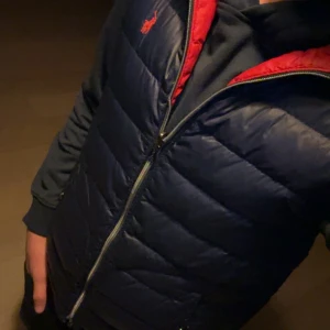 Mörkblå väst Polo Ralph Lauren - Mörkblå väst från polo Ralph lauren, där nere i knäppet kan de åka upp som man ser på bilden men man kan bara vika in den lite så syns de inte. Västen är i bra skick och har inga skado förutom där nere på dragkedjan. Man kan ha den som både röd väst och blå väst, därav sitter lappen i fickan.