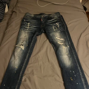 Dsquared2 blå distressed jeans herr - Säljer ett par blå Dsquared2 jeans med slitna detaljer, färgstänk och tydlig tvättad effekt. Jeansen har raka ben, klassisk femficksdesign och läderpatch bak i midjan. Materialet är bomull och de har en cool, streetig vibe.