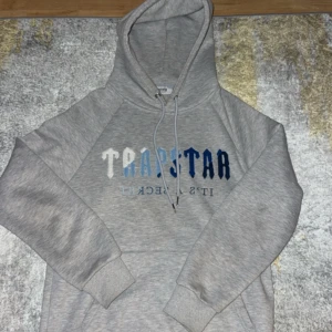 Grå hoodie Trapstar med blå text - Säljer en grå hoodie från Trapstar med fet logga i vitt och blått framtill. Tröjan har huva med snörning och är tillverkad i mjuk bomullsblandning. Perfekt för dig som gillar streetwear vibes och vill sticka ut med snygg branding.