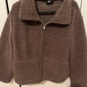 Brun teddyjacka från 157, storlek S - Mysig brun teddyjacka från 157 i storlek S. Jackan har en hög krage, dragkedja framtill och två stora fickor. Perfekt för kyliga dagar och ger en chill vibe med sin oversized look och fluffiga material. Har endast använts en gång. 