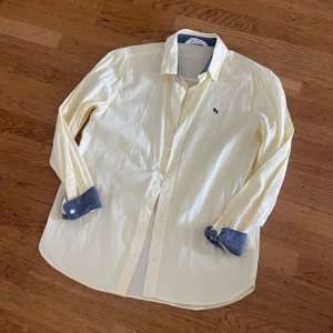 Ljusgul skjorta med blå detaljer - Fräsch ljusgul skjorta från Abercrombie & Fitch med blå kontrast på insidan av krage och manschetter. Klassisk passform, långärmad och med knappar framtill. Liten broderad logga på bröstet. Perfekt för dig som gillar stilrena och coola detaljer.