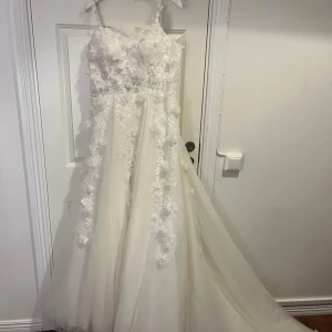 Vit bröllopsklänning från Ivory & Grace - Magisk krämvit långklänning från Ivory & Grace med tunna axelband, 3D-blommor, pärlor och spetsdetaljer. Klänningen har vacker släp, snörning i ryggen och är gjord i flera lager tyll och spets. Perfekt för dig som vill ha en romantisk och lyxig look. (Klänningen har använts endast 1 gång). Nypris 17000 kr. Storlek är lite oklar då den är anpassat sydd för mig. Jag är 162 cm lång och vid intresse kan jag skicka bild på hur den såg ut på mig. 