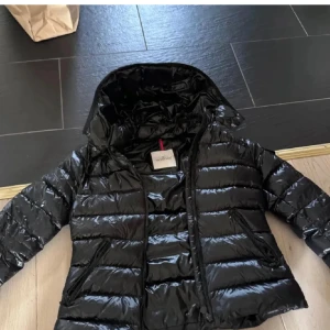 Svart Moncler pufferjacka med huva - Säljer en svart pufferjacka från Moncler med glansig finish och huva. Jackan har dragkedja framtill, två sidofickor med dragkedja och Moncler-logga på ärmen. Perfekt för kalla dagar och har en riktigt snygg streetstil.