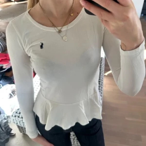 Vit långärmad topp från Ralph Lauren - Säljer en vit långärmad topp från Ralph Lauren med volangkant nedtill och den klassiska lilla loggan broderad på bröstet. Toppen har rund halsringning och är figurnära i passformen. Perfekt att styla med jeans eller kjol för en clean look.
