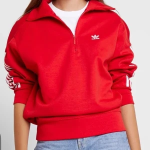 Röd Adidas Originals half zip sweatshirt - Röd Adidas sweatshirt med klassiska vita ränder längs ärmarna och broderad logga på bröstet och ärmen. Tröjan har half zip, hög krage och är tillverkad i mjuk bomullsblandning. Perfekt för en sportig och avslappnad stil.                                 Sparsamt använd. Strl 34.