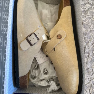 Birkenstock Boston beige mocka - Birkenstock Boston i beige mocka med justerbart spänne i metall och klassisk korksula. Skorna har en platt svart yttersula med mönster för extra grepp. Perfekt slip-on modell med rund tå och stilren design som funkar året runt.