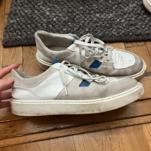 Sneaky Steve vita och grå sneakers - Snygga sneakers från Sneaky Steve i vitt med blå detaljer. Ovandelen är i mocka och skinn, vilket ger en schysst kontrast mellan materialen. Perfekta för dig som gillar stilrena men ändå unika sneakers. Knappt använda. 