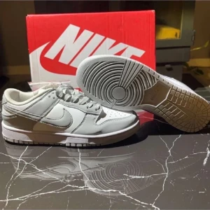 Nike Dunk Low vit och grå - Nike Dunk Low sneakers i en clean vit och ljusgrå färgkombination. Skorna har klassisk låg siluett, snörning och rund tå. Tillverkade i skinn och syntetmaterial med vit sula och grå detaljer. Perfekta för dig som gillar stilrena och trendiga sneakers.