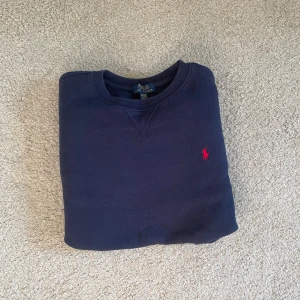 Mörkblå sweatshirt från Polo Ralph Lauren - Klassisk mörkblå sweatshirt från Polo Ralph Lauren med röd broderad logga på bröstet. Tröjan har rund halsringning, långa ärmar och är tillverkad i mjuk bomullsmix med fleeceinsida för extra komfort.