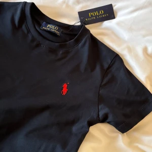Svart t-shirt från Polo Ralph Lauren - Svart t-shirt från Polo Ralph Lauren med klassisk rund hals och korta ärmar. Ikoniska röda broderade loggan på bröstet. Tillverkad i mjuk bomull för en skön och stilren look. Perfekt för dig som gillar clean och tidlös stil.