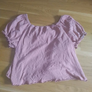 Rosa blus med puffärm från KappAhl - Säljer en rosa blus från KappAhl i storlek 158/164. Blusen har puffiga korta ärmar med rynkade kanter och en rundad halsringning. Materialet är mjukt och känns som bomull, perfekt för dig som gillar en söt och enkel stil.