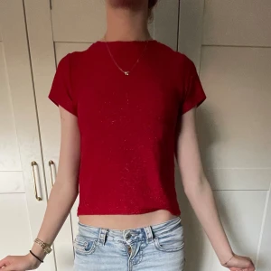 Röd croppad topp från KappAhl - Snygg röd croppad topp från KappAhl med korta ärmar och enkel rund halsringning. Toppens material har en lätt glittrig yta som ger extra vibe. Perfekt att matcha med jeans eller kjol för en cool look.
