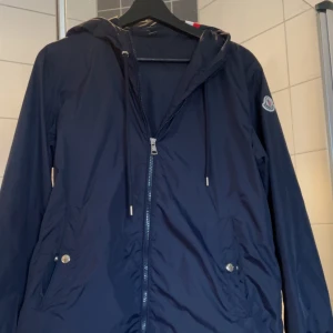 Moncler Jacka - Mörkblå jacka från Moncler,har vit-röda ränder som en detalj på luvan.Storleken passar 160-175.för mer frågor och funderingar kontakta mig 😃