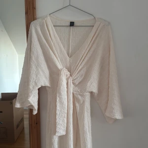 Lång ljus omlottklänning från SHEIN - Lång ljusbeige omlottklänning från SHEIN med v-ringning och knyt framtill. Klänningen har vida trekvartsärmar och är gjord i ett strukturerat, lätt material som ger en luftig känsla. Perfekt för sommarens varma dagar.