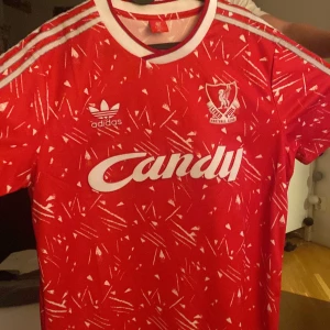 Liverpool retrotröja Adidas XL röd - Retro Liverpool FC fotbollströja från Adidas i rött med vita detaljer och ikoniskt Candy-tryck på bröstet. Tröjan har korta ärmar, klassiska Adidas-ränder på axlarna och ett unikt abstrakt mönster. Tillverkad i lätt och glansigt material, perfekt för fans.