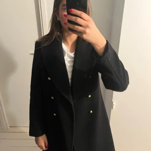 Svart kappa från Club Monaco - Säljer en svart lång kappa från Club Monaco med dubbelknäppning och guldiga knappar. Klassisk krage och rak passform, perfekt för dig som vill ha en stilren och tidlös look. Gjord i ullblandning och har långa ärmar.