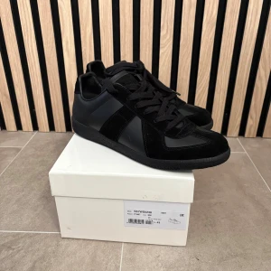 Svarta Maison Margiela sneakers - Maison Margiela sneakers i helsvart med snygga paneler i skinn och mocka. Klassisk låg modell med rund tå, platt sula och snörning. Diskret branding på insidan och yttersulan har coolt mönster. Perfekt för dig som gillar minimalistisk och stilren vibe. Bild 5 visar en skavank.SÄLJS PÅ GRUD AV DE ÄR FÖR SMÅ!!