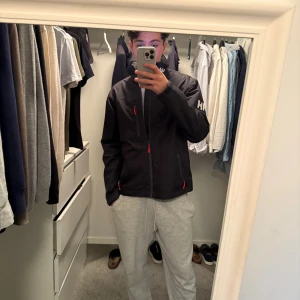 Svart vindjacka från Helly Hansen - Svart vindjacka från Helly Hansen med hög krage och vita logodetaljer på ärm och krage. Jackan har röda dragkedjor på fickor och framtill, samt en clean och sportig look. Perfekt för dig som gillar stilren och funktionell design.