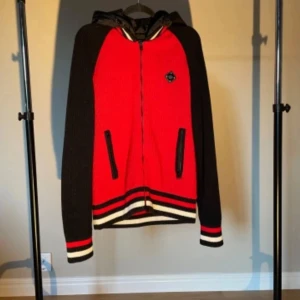 Cardigan jacka från J.Lindeberg - Snygg röd och svart jacka från J.Lindeberg med dragkedja och huva. Jackan har ett ribbat mönster och randiga detaljer vid ärmslut och nederkant. Perfekt för en cool och avslappnad stil. Nypris 2999