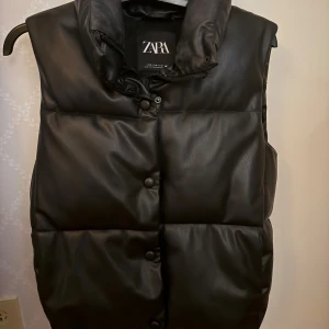 Svart dunväst i skinnimitation från Zara - Snygg svart dunväst från Zara i skinnimitation med hög krage, tryckknappar och dragkedja. Västen är quiltad och har praktiska fickor på sidorna. Perfekt för lager-på-lager och ger en cool vibe till din outfit.