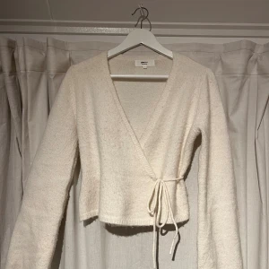 Vit omlottkofta från Gina Tricot - Mysig vit omlottkofta från Gina Tricot i ullblandning. Koftan har lång ärm, knytband i midjan och en croppad passform. Perfekt för lager-på-lager och chill dagar. Enkel och stilren design som funkar till allt.