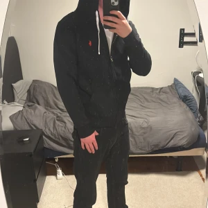 Svart hoodie från Polo Ralph Lauren - Svart hoodie från Polo Ralph Lauren med dragkedja, vit snörning i huvan och röd broderad logga på bröstet. Klassisk passform och mjukt bomullsmaterial, perfekt för en chill och stilren look.