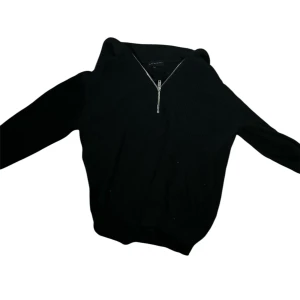Svart stickad hoodie Brand Island S - Svart stickad hoodie från Brand Island i storlek S. Tröjan har en ribbad struktur, huva och en snygg half zip-dragkedja framtill. Perfekt för dig som gillar en enkel men stilren look med lite edge.