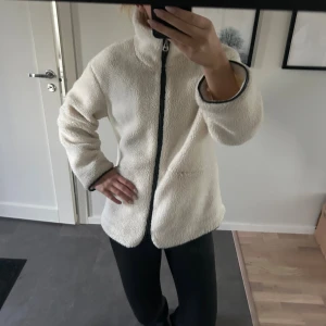 Vit teddyjacka från Vero Moda - Mysig vit teddyjacka från Vero Moda med hög krage och hel dragkedja framtill. Jackan har svarta detaljer längs dragkedjan och ärmarna, samt två fickor på sidorna. Storlek s men är stor i storleken och bra skick.