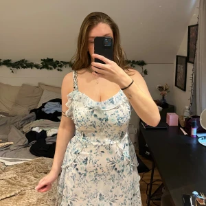 Blommig kort klänning från Trendyol - Superfin vit kort klänning med grön/blått blommigt mönster från Trendyol. Klänningen har smala axelband, volangdetaljer vid halsringningen och längs kjolen samt en lätt v-ringad front. Perfekt för sommarens alla tillfällen!