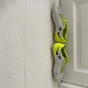 Nike fotbollsskor vit och neon - Säljer ett par grymma Nike fotbollsskor i vit och neon-gul färg med svart swoosh på sidan. Skorna har snörning, platt sula med dobbar och rund tå. Materialet är syntet och skinn, med coola detaljer som svensk flagga och texten 'DJ6' på sidan.