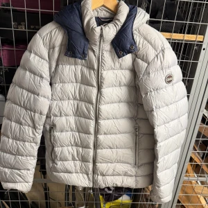 Ljusgrå dunjacka från Colmar - Säljer en ljusgrå dunjacka från Colmar med marinblå detaljer vid huvan. Jackan har horisontella quiltade sömmar, två fickor med dragkedja och en snygg logotyp på ärmen. Perfekt för kalla dagar med sin fluffiga och isolerande design.