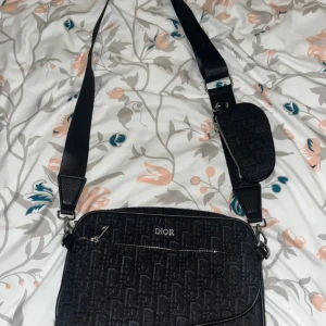 Dior väska - Snygg svart axelväska från Dior med tydligt monogrammönster och silverfärgade dragkedjor. Väskan har bred axelrem och en extra liten ficka med dragkedja på remmen. Perfekt för dig som vill ha en stilren och trendig väska med plats för det viktigaste.