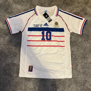 HELT NY Frankrike retro VM 98 Zidane - HELT NY! RETRO Frankrike fotbollströja från VM 1998 med blå och röda detaljer, nummer 10 och Zidane på ryggen. Adidas-logga, FFF-märke och broderat Frankrike märke på bröstet. Tillverkad i lätt och ventilerande material, perfekt för RIKTIGA fotbollsfans!
