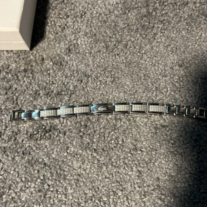 Silverfärgat armband från Lacoste - Stilrent armband från Lacoste i silverfärgad metall med rektangulära länkar och diskret rutmönster. På en av länkarna syns den klassiska Lacoste-krokodilen. Kommer med original ask och vit tygpåse med logga. Perfekt accessoar för dig som gillar minimalistisk design.