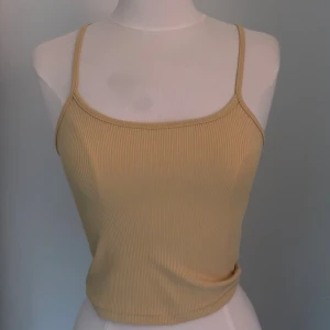 Gul ribbad linnetopp med smala band - Säljer en gul ribbad linnetopp med smala axelband och croppad passform. Toppen har en enkel och stilren design som är perfekt för varma dagar. Materialet är mjukt och stretchigt för en bekväm känsla.