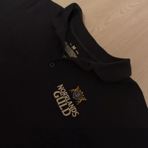 Mörkblå pikétröja Norrlands Guld  - Mörkblå pikétröja med klassisk krage och två knappar framtill. Broderad Norrlands Guld-logga i vitt, blått och guld på bröstet. Storlek M. Skick 8/10. Skriv vid minsta fundering!