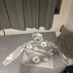Grå hoodie med vita blommor Denim Tears - Säljer en grå hoodie från Denim Tears i storlek S. Tröjan har vita blommönster över hela, huva och en stor magficka. Perfekt för dig som gillar streetwear och vill sticka ut med en unik design.