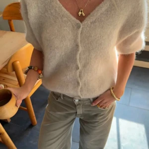 Beige fluffig kofta med knappar - Supermjuk och fluffig beige kofta med v-ringning och knappar framtill. Koftan har kortare ärmar och en avslappnad passform, perfekt för lager på lager. Den stickade känslan ger en mysig vibe och passar till både jeans och kjol.