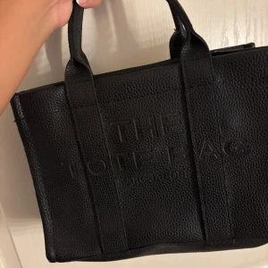 The tote bag från Marc Jacobs Ej äkta  - Säljer en svart handväska från Marc Jacobs, The Tote Bag. Väskan är tillverkad i strukturerat skinn med präglad logga på framsidan. Den har två rejäla handtag och en boxig form som gör den både snygg och praktisk. Perfekt för dig som gillar stilrena accessoarer.