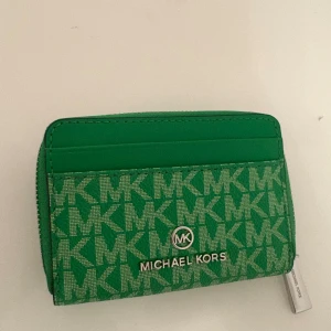 Grön MK plånbok med logomönster - Snygg grön plånbok från Michael Kors med MK-logomönster över hela ytan. Plånboken har flera kortfack, silverfärgad dragkedja och metallogo framtill. Materialet är slitstark syntet och designen är kompakt och praktisk för vardagsbruk.