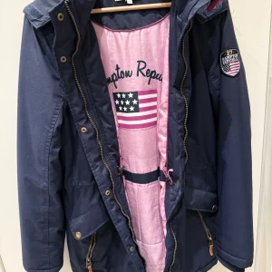 Marinblå parkas från Hampton Republic - Snygg marinblå parkas från Hampton Republic med rosa foder och patch på ärmen. Jackan har huva med beige fuskpäls, dragkedja och knappar framtill samt praktiska fickor. Perfekt för kyliga dagar och har en sportig, amerikansk vibe. Storlek: 170 