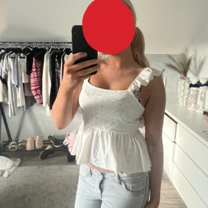 Vit volangblus med broderi - Söt vit blus med volangaxelband och broderade detaljer. Toppen har en peplumformad nederdel och är ärmlös, perfekt för ljusa dagar. Luftigt bomullsmaterial och romantisk känsla.