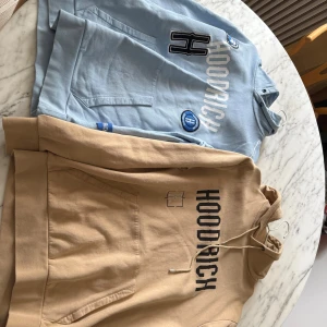Två Hoodrich hoodies i blått och beige - Säljer två snygga Hoodrich hoodies, en i ljusblått med broderade patches och tryck, och en i beige med svart tryck. Båda har huva med snörning och stor magficka. Perfekta för en chill och street-inspirerad look.