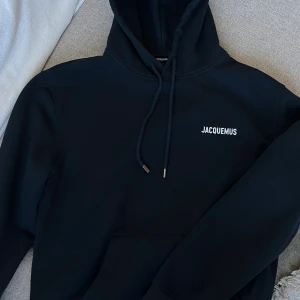 Mörkblå hoodie från Jacquemus - Säljer en mörkblå hoodie från Jacquemus med vit logga på bröstet. Tröjan har huva med snörning och metalltips med Jacquemus-gravyr. Klassisk känguruficka framtill och mjukt material som känns riktigt skönt. Den är i nyskick endast testad! Nypris 3200kr. Strlk XL men passar L