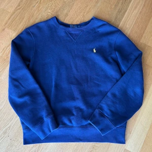 Ralph Lauren tröja - Klassisk blå sweatshirt från Polo Ralph Lauren med rund halsringning och broderad gul logga på bröstet. Tröjan har ribbade muddar vid ärmslut och nederkant, perfekt för en stilren och avslappnad look.