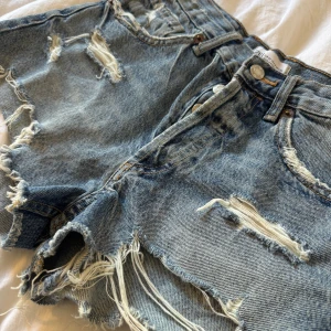 Blå slitna jeansshorts från Zara - Säljer ett par blåa högmidjade jeansshorts från Zara med råa, fransiga kanter och flera slitningar framtill. Klassisk femficksmodell med knappgylf och bälteshällor. Perfekta för en avslappnad och trendig look i sommar.